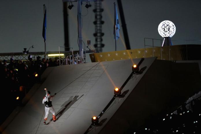 A man carrying the torch on his back climbing a slope using a rope.