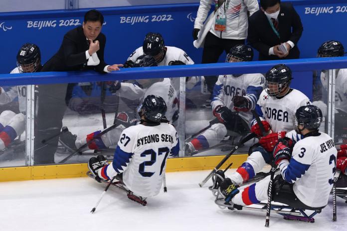 A male coach wearing a black suit speaks to Para ice hockey players on the rink. 