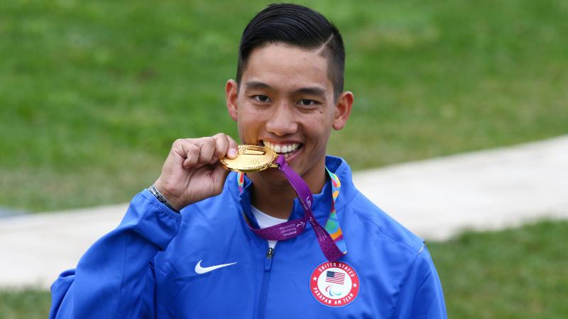 a male Para shooter biting a gold medal and smiling