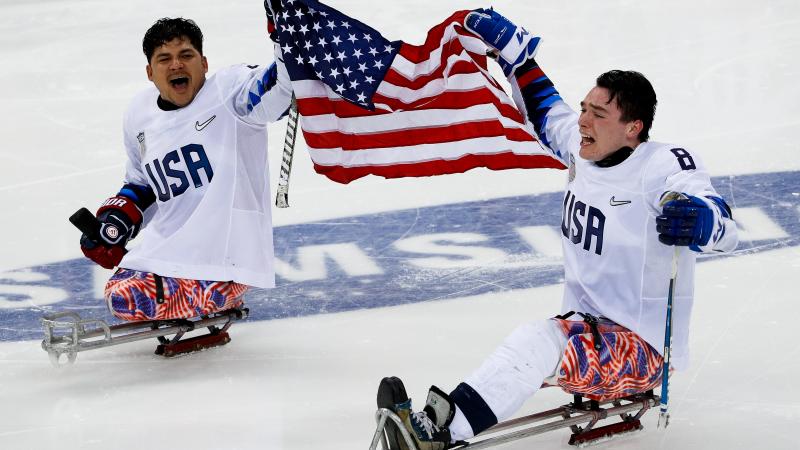 two male US Para ice hockey players on the ice holding a USA flag