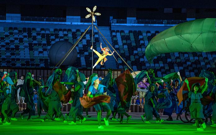 Performers in green and blue outfits dance, with one on supports in the air, with one of three large inflatable Agitos flying into the scene