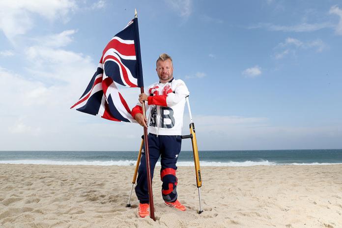 Sir Lee Pearson, supported by crutches, holds up a British flag on the beach in Rio de Janeiro.