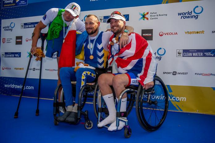Two male athletes using wheelchairs and one male athlete poses for a photograph after receiving their medals at the 2022 World Rowing Championships.