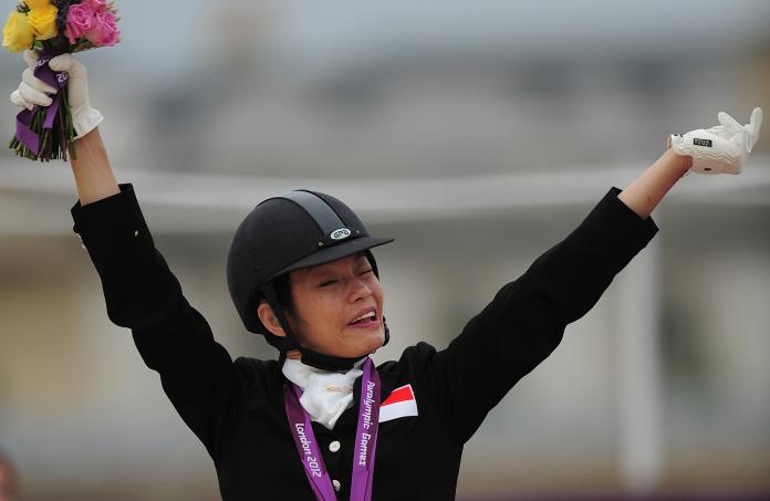 A female Para equestrian rider with a medal around her neck waves a flower bouquet at an awards ceremony.