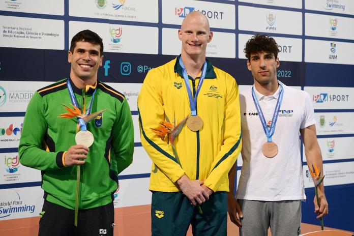 Three male swimmers in their national kit smile as they stand on the podium with medals around their necks.