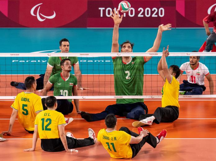 A tall sitting volleyball player returns a serve over the net as his teammates and opponents follow the move with their eyes.