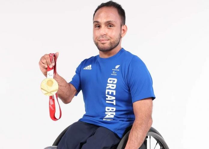 A male athlete in a blue Great Britain T-shirt sits on a wheelchair and holds a Paralympic gold medal with his right hand
