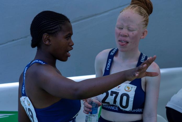 Two female sprinters talk on the sidelines of a stadium, one raising her hand and spreading the fingers on her palm as she speaks.