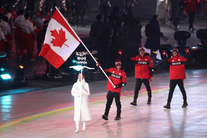 A male athlete carries the Canadian flag at the Opening Ceremony followed by two members of the Canadian delegation.