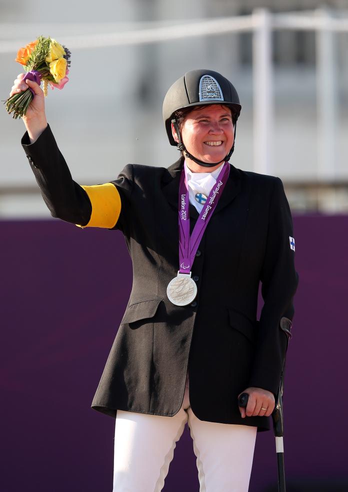 A female Para equestrian rider waves her bouquet as she stands on the podium with a medal around her neck and a crutch in her left hand.