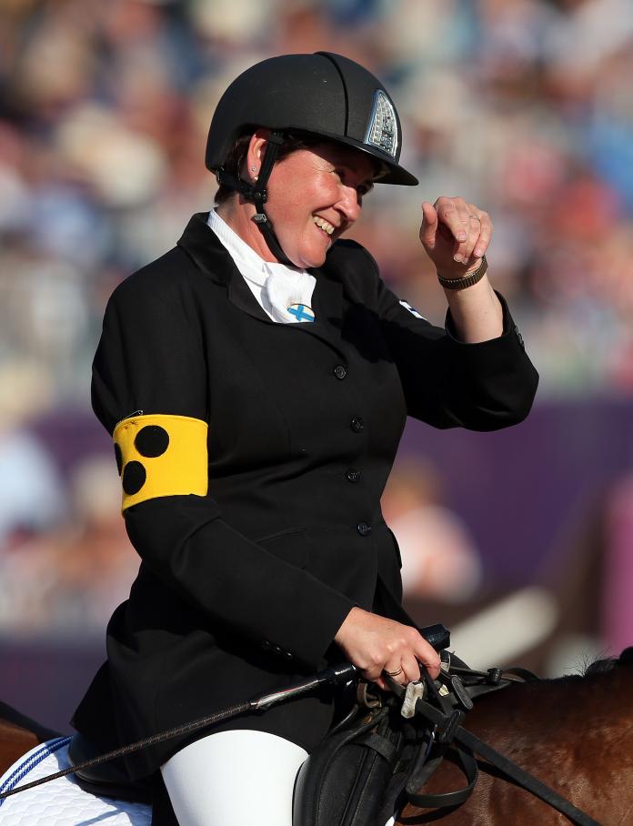 A female Para equestrian rider, seated on a horse, smiles as she takes her left hand away from her helmet while holding the reins with her right hand.
