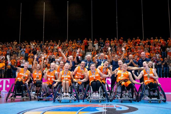 About 20 female wheelchair basketball players and officials celebrate in front of a crowd