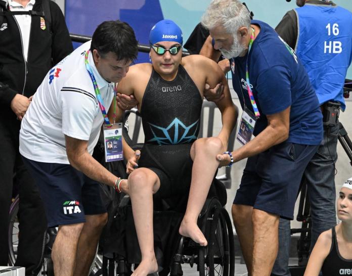 Two male coaches lift a female athlete from her wheelchair.