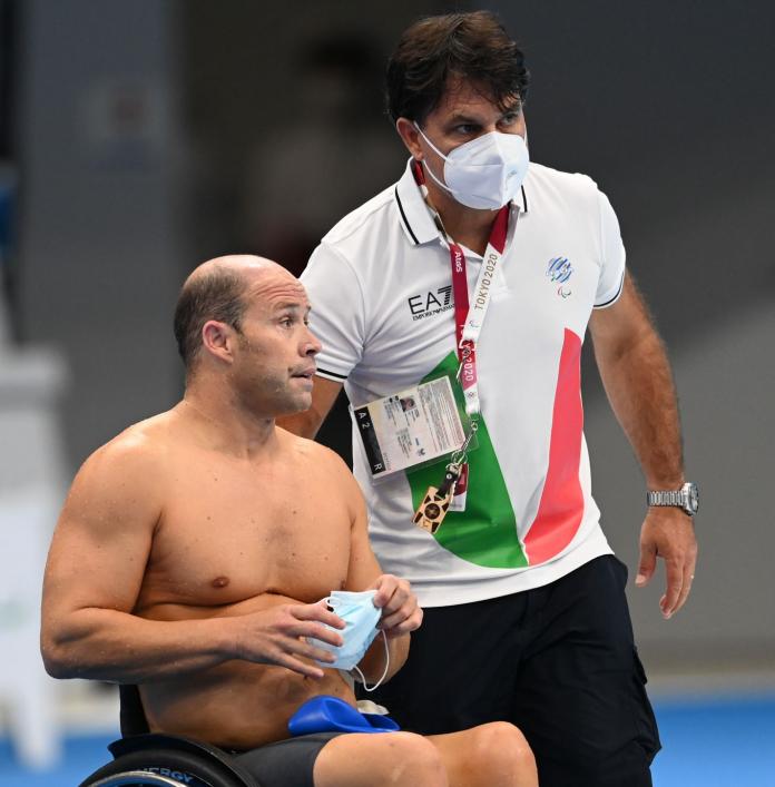 A male coach wearing a mask stands next to a male Para swimmer who is using a wheelchair.