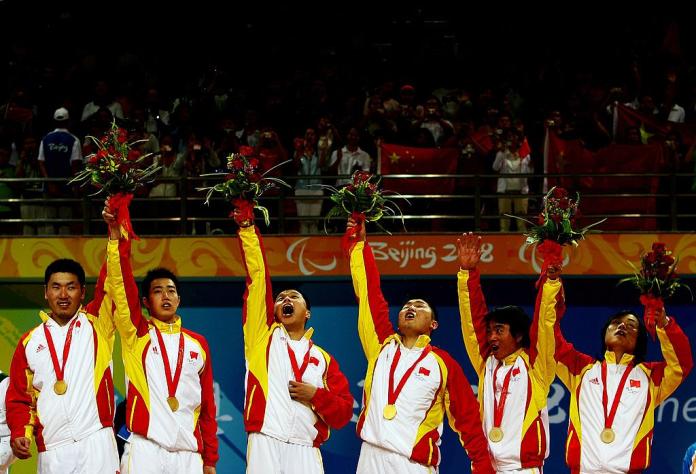 Six male goalball players celebrate on the podium by raising their hands.