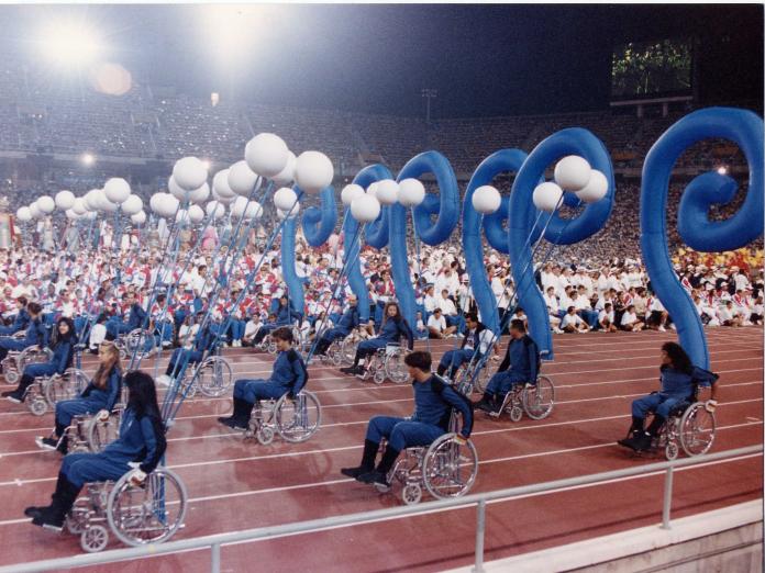 People in wheelchairs roll in wheelchairs on an athletics track with balloons fixed to their chairs. 