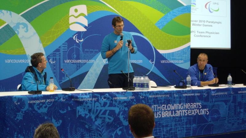 A man standing up and speaking in front of a Vancouver 2010 backdrop
