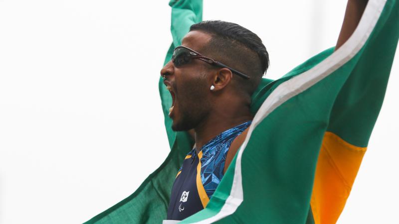 a male Para athlete cheers and holds up the Brazil flag