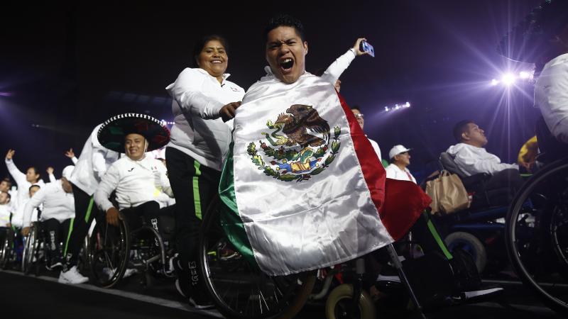 a male wheelchair athlete holds up a Mexican flag and cheers