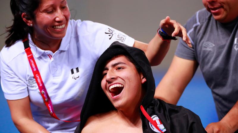 a male Para swimmer smiles as his assistants put his jacket on