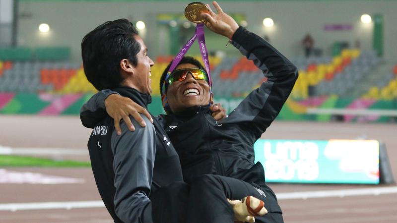 a male vision impaired runner being carried by his guide while waving his medal