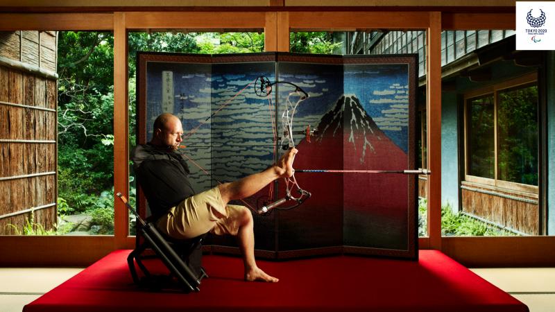 Armless archer sits down holding bow and arrow with foot with Japanese backdrop