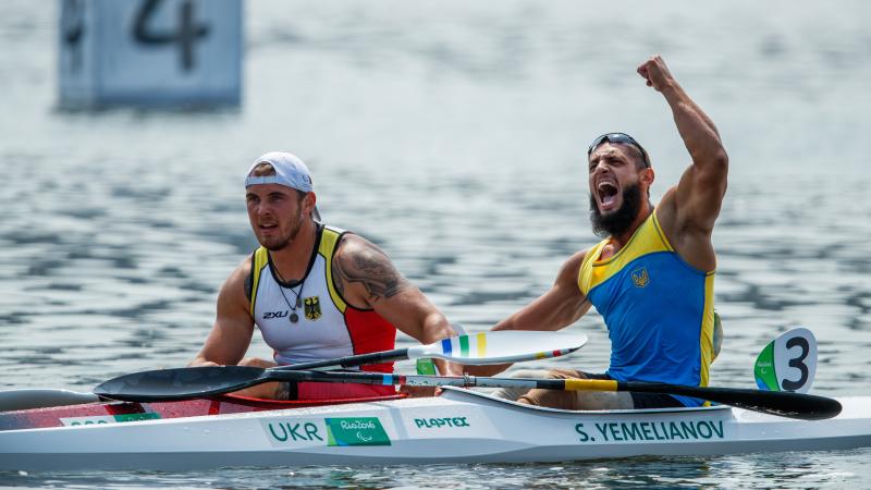 two male Para canoeists side by side in their boats with one punching the air in celebration