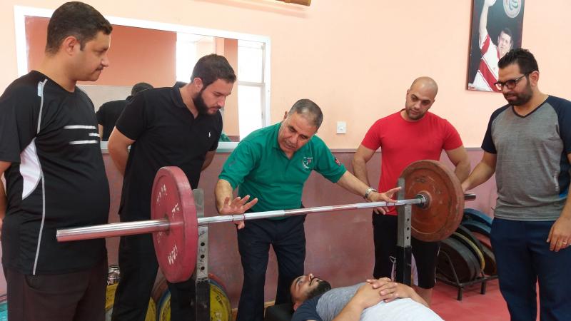 a group of officials standing round an athlete lying on a powerlifting bench