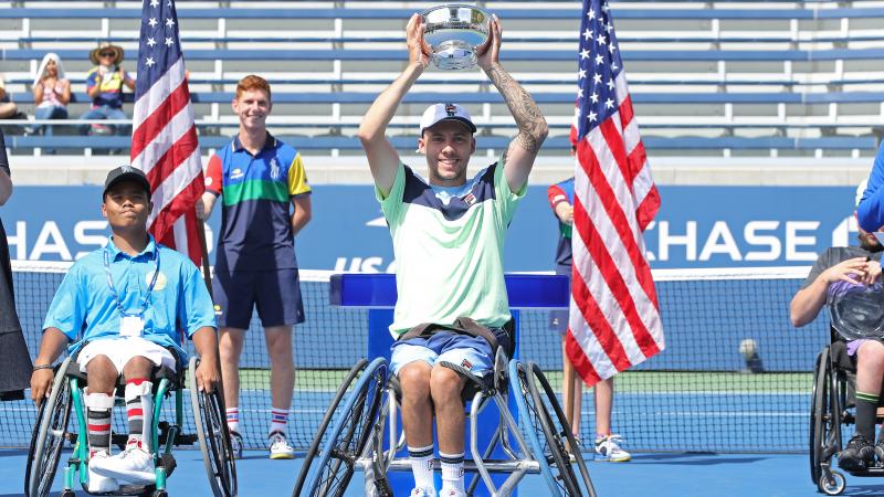 a male wheelchair tennis player holds up a trophy