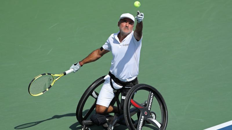 a male wheelchair tennis player throws the ball up to serve