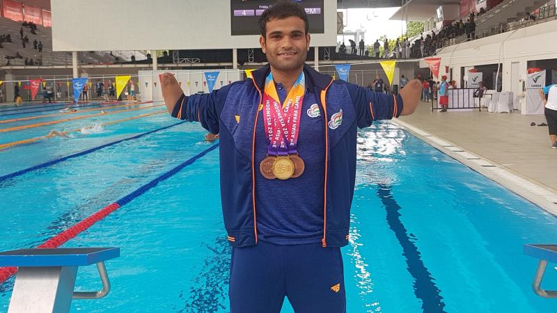 a male Para swimmer standing in front of a swimming pool with medals round his neck