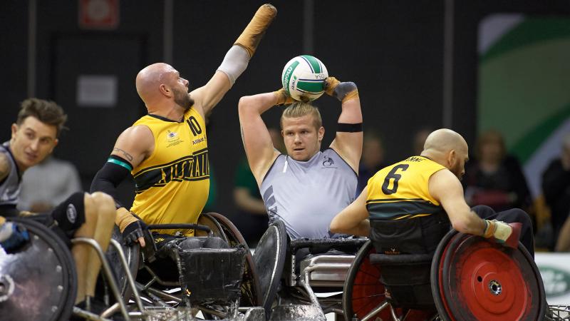 New Zealand male wheelchair rugby player holds ball over head while Australian defenders try surround him
