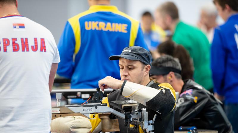 A man loading his rifle during a shooting competition with other competitors and a man standing next to him