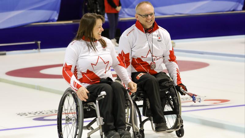 Man and woman in wheelchairs smile on curling ice sheet