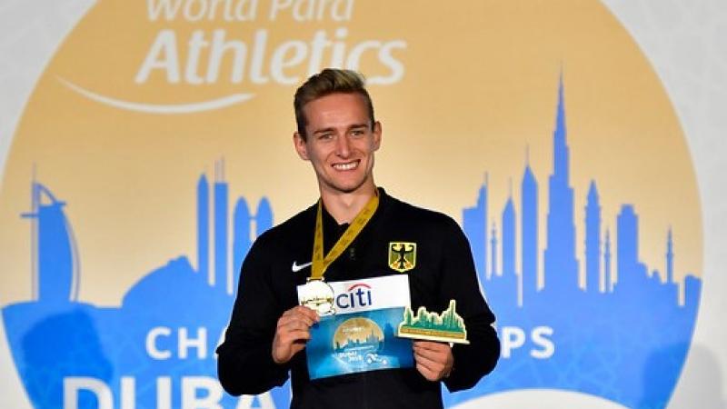 Man smiles on podium holding gold medal and prize
