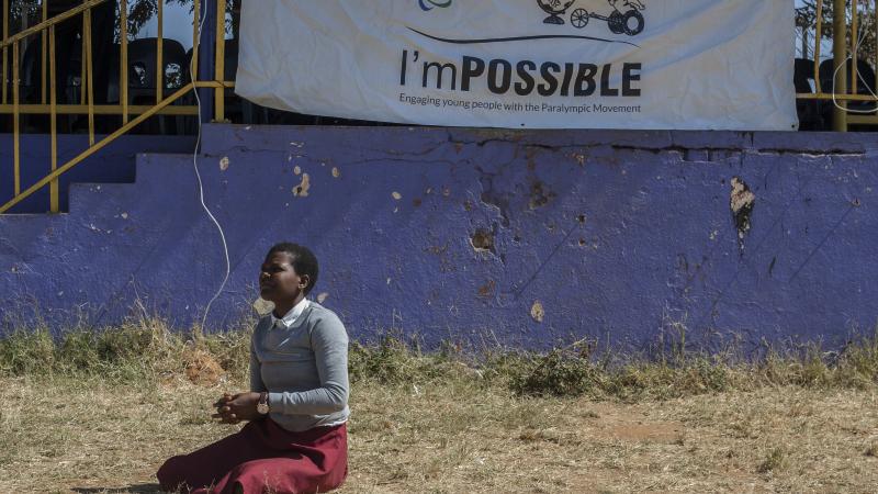 Student sits on the grass during an I'mPOSSIBLE activity in Malawi, with a flag of the programme in the background