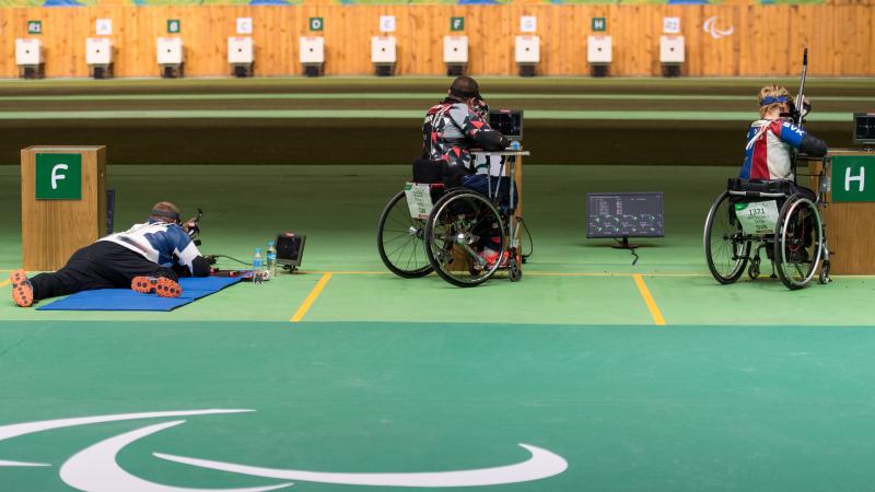 A man on the floor and two persons in wheelchairs with rifles aiming at targets in a shooting Para sport competition