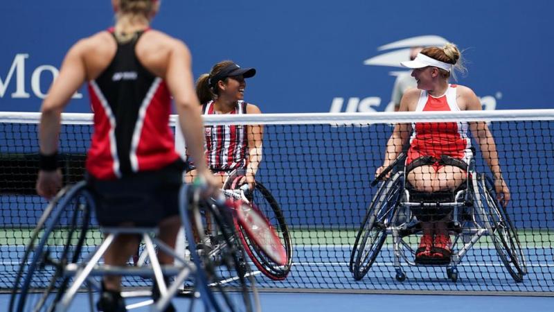 Two female wheelchair tennis doubles partners smile at each other after winning