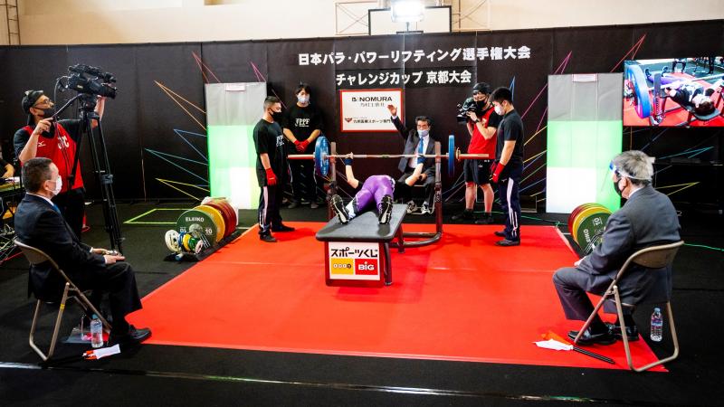 A person on a bench press during a Para powerlifting competition observed by three judges, three loaders and two cameramen