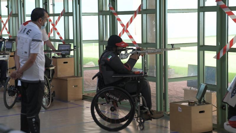 A man standing behind a man in a wheelchair shooting with a rifle in a shooting range