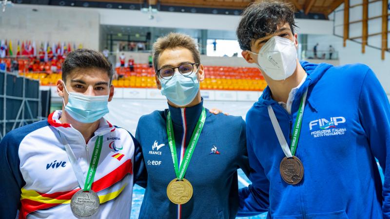 Three men together with their medals in front of a swimming pool