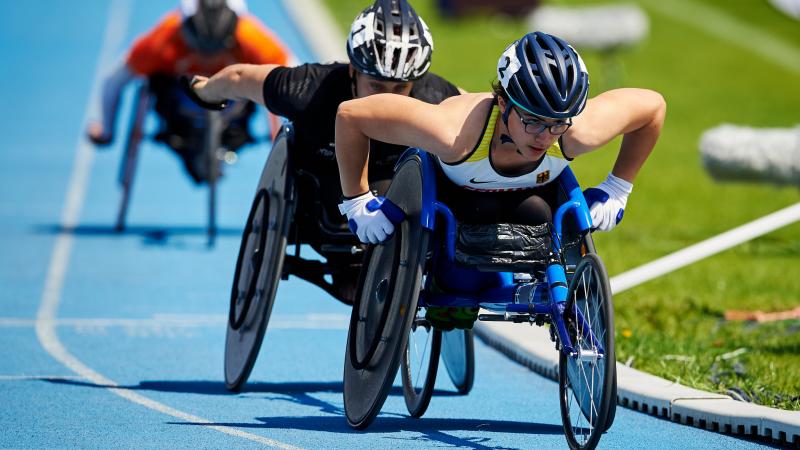 A woman in a racing wheelchair being followed by two athletes during a competition
