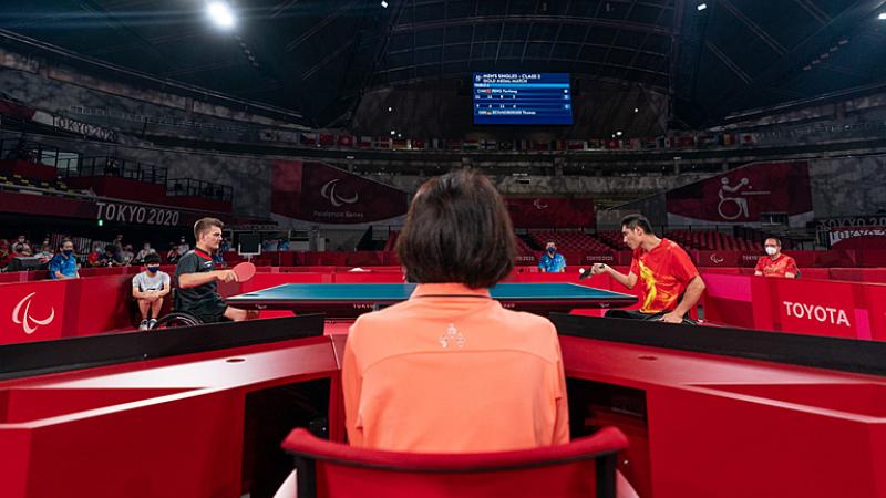 Two male wheelchair table tennis players playing with judge looking on
