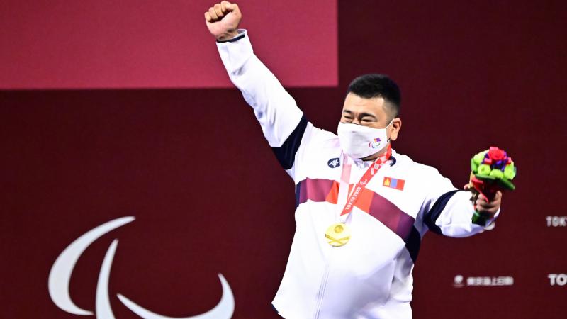 A man holding his right hand high with a gold medal around his neck and a mask on his face.