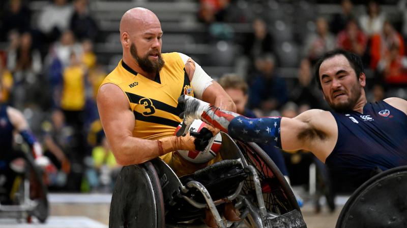 A male wheelchair rugby player wearing a yellow jersey holds onto a ball, while another athlete wearing a blue jersey tries to grab it