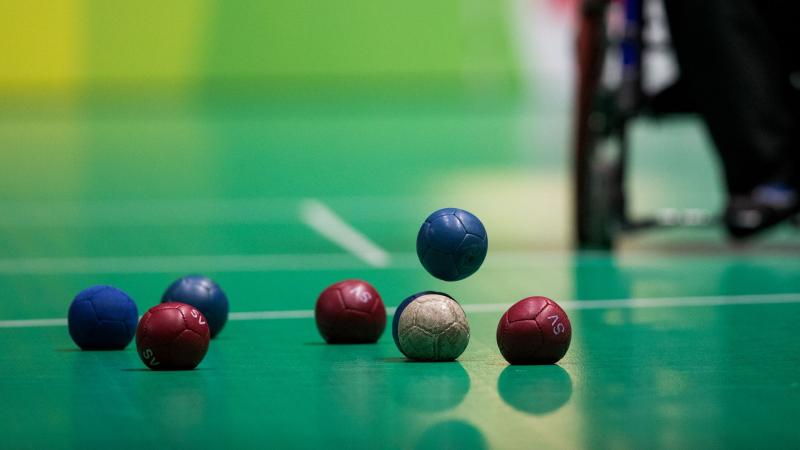 Seven boccia balls lying on the playing ground while one blue ball is falling down, about to touch the ground.