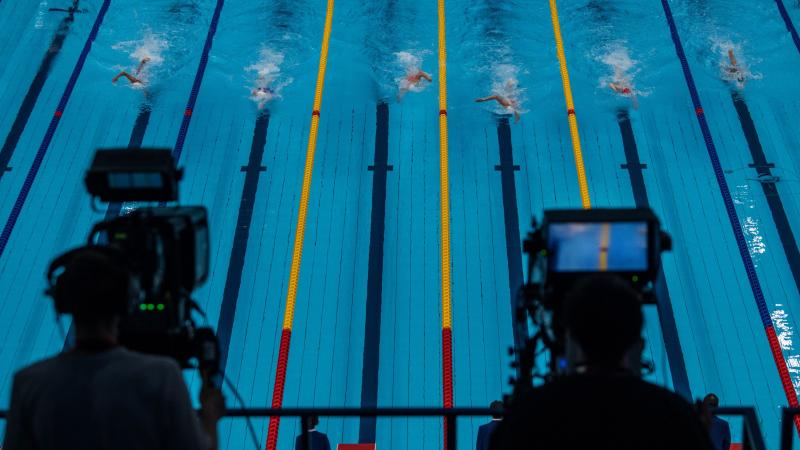 A look at a competition pool in Tokyo, with six swimmers in the lanes, through two cameras that are filming the race.