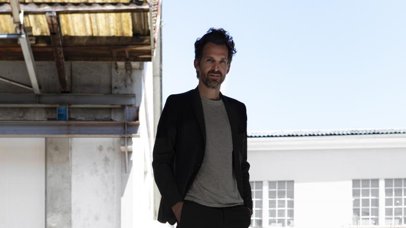 A man in a T-shirt and black suit poses with the backdrop of a slightly dilapidated house.