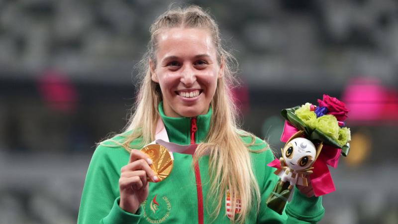 A female athlete smiles for a photograph while holding a gold medal with her right hand and a bouquet with her left hand.