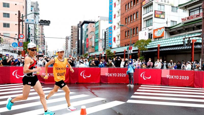 A male runner and his guide compete in the marathon at Tokyo 2020. People are taking photos as they compete.
